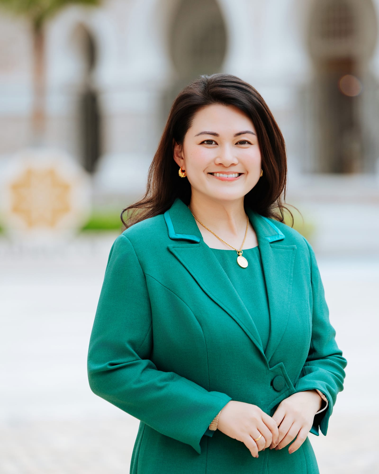 Chan Quin Er in a jade-green suit, smiling, in a courtyard of white arches.