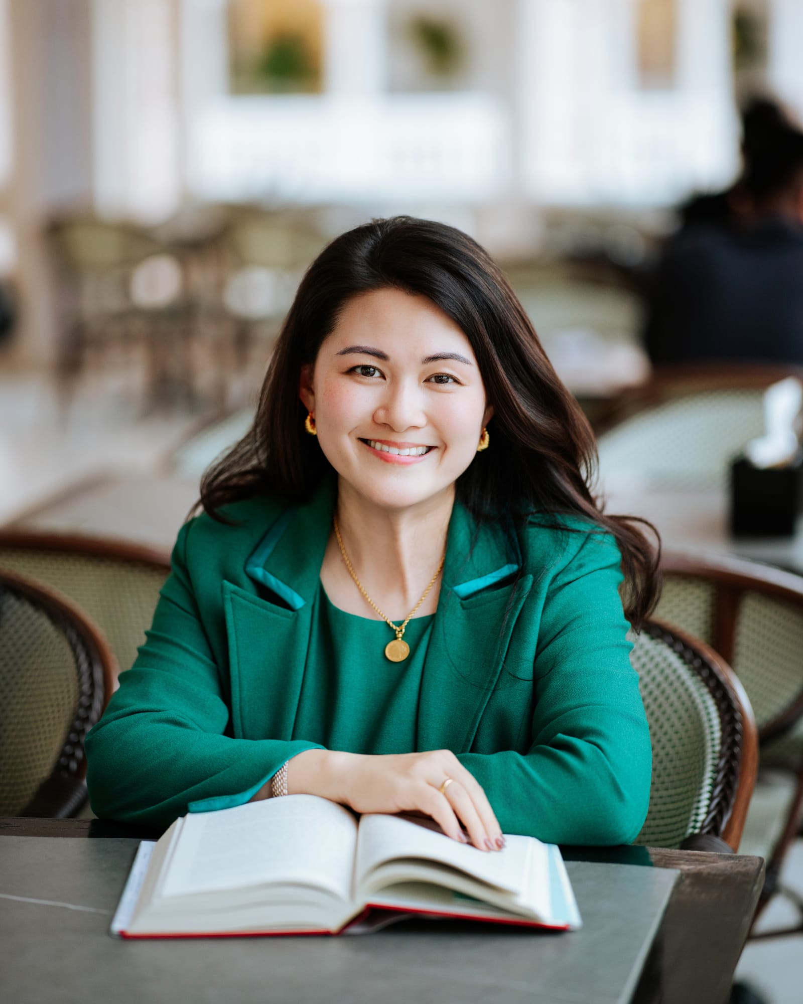 Chan Quin Er in a jade-green suit at a café table, an open book in front of her, smiling at the camera.
