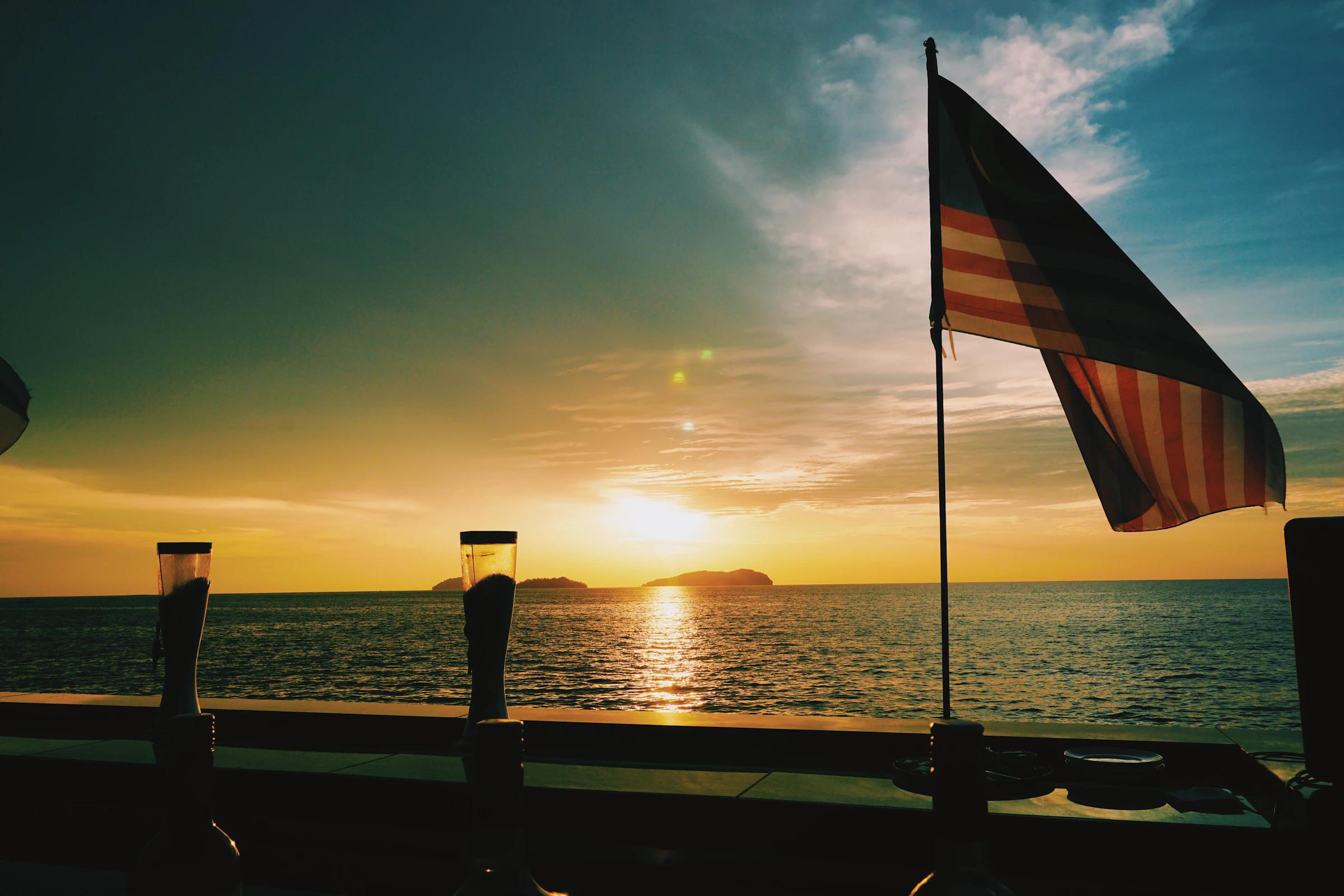 The Jalur Gemilang in silhouette against a sunset over the sea.